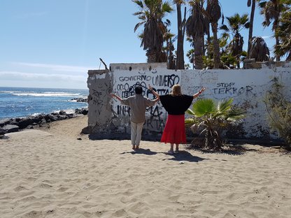Ausstellungseinladung: 2 Menschen mit ausgebreiteten Armen vor Meer und gesprayter Mauer. Fotoprojekt Edith Tarr